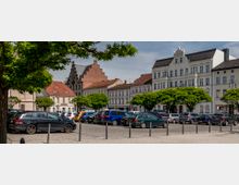 Ein öffentlicher Platz mit parkenden Autos, umgeben von historischen Gebäuden mit teils verzierten Fassaden und einem markanten Gebäude mit rotem Treppengiebel im Hintergrund. Im Vordergrund stehen Bäume mit grünen Kronen, die den Platz säumen.