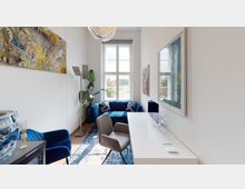 Minimalistisch eingerichtetes Büro oder Arbeitszimmer mit einem großen Fenster, das viel Tageslicht hineinlässt. Im Raum befinden sich ein Schreibtisch mit Computer, ein grauer Stuhl, ein blaues Sofa, moderne Kunstwerke an den Wänden und Pflanzen als Dekoration.
