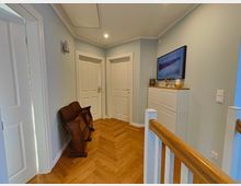 Ein hell beleuchteter Flur mit Parkettboden in Fischgrätenmuster, zwei weißen Türen, einer weißen Kommode mit Dekoration und einem gerahmten Bild an der Wand. Im Vordergrund ein weißes Treppengeländer mit Holzhandlauf und an der linken Wand zwei zusammengeklappte Holzbänke.