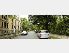 Straße mit Kopfsteinpflaster, links ein mehrstöckiges historisches Gebäude mit sandfarbener Fassade und großen Fenstern, rechts dichter Baumbestand; parkende Autos entlang beider Straßenseiten.