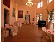 Ein eleganter Veranstaltungsraum mit weiß gedeckten Tischen, Holzfußboden, hohen Decken und großen Fenstern. Der Raum ist geschmückt mit Kunstwerken an den Wänden, einem Kronleuchter und Topfpflanzen, sowie einem schwarzen Klavier in der Ecke.