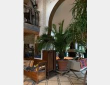 Ein gemütlicher Innenraum mit mediterranem Stil, der durch viele große Topfpflanzen, eine hölzerne Sitzbank mit bunten Kissen, einen Kamin und rustikale Möbel definiert wird. Sichtbare Steinwände, ein großer Bogen und ein gefliester Boden mit dekorativem Muster unterstreichen das Ambiente.