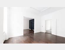 Heller, leerer Wohnraum mit weiß gestrichenen Wänden, dunklem Holzboden und großen Türöffnungen, die zu weiteren Räumen führen, von denen einer eine dunkelgraue Akzentwand aufweist.