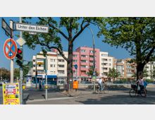 Straßenkreuzung mit Wohngebäuden im Hintergrund, darunter mehrstöckige, bunt angestrichene Gebäude mit Geschäften im Erdgeschoss. Im Vordergrund ein Straßenschild mit der Aufschrift „Unter den Eichen“, Bäume, ein Fahrradwegzeichen sowie eine Ampel; einige Personen fahren mit Fahrrädern.