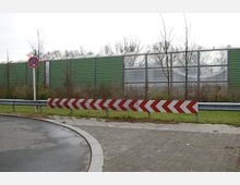 Eine Straße mit einer Kurve, abgesichert durch eine Leitplanke und eine rot-weiß gestreifte Begrenzung; im Hintergrund eine grüne Lärmschutzwand mit durchsichtigem Abschnitt sowie kahle Bäume. Ein Verkehrszeichen zeigt ein "Haltverbot".