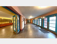 Ein langer, innenliegender Flur mit blau gerahmten Glastüren auf der rechten Seite und einer gelb-rosa Wandgestaltung. Links befindet sich eine Garderobe mit hölzernen Ablagen und Kleiderhaken, der Boden besteht aus grauen Fliesen.