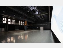 Großer, moderner Veranstaltungsraum mit Betonboden, dunklen Wänden und einer hohen Decke mit sichtbaren Stahlträgern und Lichttechnik. Links Fenster und eine Empore mit Geländern, rechts eine große, beleuchtete Leinwand.