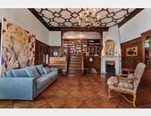 Wohnzimmer mit Holzwänden und aufwändiger Kassettendecke, ausgestattet mit einem hellblauen Sofa, einem Sessel, Kamin und Klavier. Über eine kurze Holztreppe gelangt man zu einer erhöhten Bibliothek mit Bücherregalen, die den Raum dominiert.