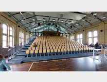 Innenaufnahme eines modernen Hörsaals in einem historischen Gebäude mit Backsteinwänden und großen Rundbogenfenstern. Die Sitzreihen bestehen aus Holzstühlen auf einer ansteigenden Tribüne, die von einem freien Dachtragwerk mit sichtbaren Metallstreben und Leuchten überdacht wird.