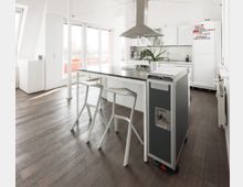 Offene, moderne Küche mit weißem Interieur, zentraler Kücheninsel mit zwei weißen Hockern und einem hängenden Dunstabzug. Heller Raum mit Holzboden, großen Fenstern, die auf einen Balkon führen, und einem grauen Rollcontainer neben der Insel.