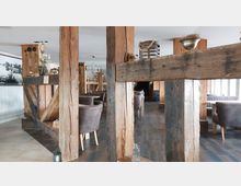 Innenraum eines rustikalen Restaurants mit sichtbaren Holzstützen und -balken. Der Raum ist mit braunen Polstersesseln, dunklem Holzboden und dekorativen Accessoires gestaltet, wie Kronleuchtern und Pflanzen.