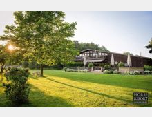 Großzügige grüne Wiese mit einem großen Baum im Vordergrund und einem zweistöckigen Gebäude aus Holz mit großen Fenstern und Terrassen im Hintergrund; Sonnenschirme und Blumenbeete umgeben die Terrasse, während die Sonne durch das Laub scheint.