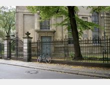 Straßenansicht eines historischen Gebäudes mit großen Fenstern und einer verzierten Metalltoranlage im Vordergrund. Ein Baum und zwei abgestellte Fahrräder vor dem Zaun ergänzen die Szene.
