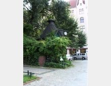 Kleiner Pavillon mit begrüntem Dach, größtenteils von Pflanzen bedeckt, mit einer analogen Uhr an der Seite. Im Hintergrund stehen Bäume sowie ein mehrstöckiges Gebäude mit heller Fassade und Fenstern.