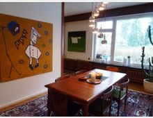 Esszimmer in einem Wohnraum mit einem Holztisch und vier Stühlen auf einem gemusterten Teppich. An der Wand hängt ein großes, modernes Gemälde mit gelbem Hintergrund, im Hintergrund steht ein großes Fenster mit Ausblick auf einen Garten, daneben befindet sich ein großer Kaktus in einem hellen Topf.