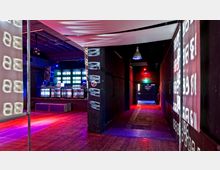 Industrieähnliches Club-Interieur mit dunklen Wänden, Holzboden und einer Bühne im Hintergrund, die von leuchtenden LED-Displays beleuchtet wird; bunte Lichter und Plakate mit Projektionen schmücken den Raum. Ein schmaler Gang führt zu einer beleuchteten Tür im hinteren Bereich.