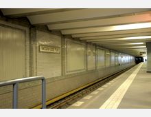 Unterirdischer U-Bahnhof „Viktoria-Luise-Platz“ mit weiß gefliesten Wänden, gelben Details entlang der Gleise und rechteckigen Architektur. Der Bahnsteig hat helle Bodenmarkierungen, und eine elektronische Anzeigetafel ist im Hintergrund sichtbar.