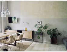 Helles Wohnzimmer mit modernem Interieur, bestehend aus einem Esstisch mit zwei Rattanstühlen und Fellauflagen auf einem hellen Teppich. An der grauen Wand befinden sich ein Holzsideboard, grüne Zimmerpflanzen, ein kleiner gerahmter Druck und ein an der Wand montierter Fernseher.