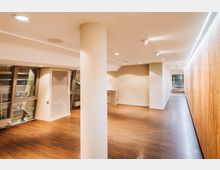 Moderner Innenraum mit Holzboden, einem weißen Säulenpfeiler in der Mitte, Deckenspots und einer Wandverkleidung aus vertikalem Holz an der rechten Seite. Große Fenster mit metallenen Rahmen an der linken Wand lassen Tageslicht herein.