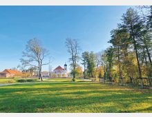 Weitläufige Parklandschaft mit einer Wiese im Vordergrund, Bäumen am Rand und einer Kirche mit rotem Dach und Zwiebelturm im Hintergrund. Links stehen Gebäude mit roten Ziegeldächern, der Himmel ist strahlend blau.