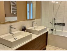 Modernes Badezimmer mit einem Doppelwaschtisch aus Holz und Marmoroberfläche, darauf zwei rechteckige Waschbecken mit Chromarmaturen. Im Hintergrund eine Dusch-Badewanne mit Glasabtrennung und weiße Wandfliesen, darüber ein Handtuchheizkörper.