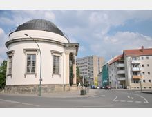 Runder, weißer klassizistischer Bau mit großen Fenstern und einem schwarzen Kuppeldach, flankiert von moderneren Gebäuden und einer Straße mit geparkten Autos; im Hintergrund teils bewölkter Himmel.