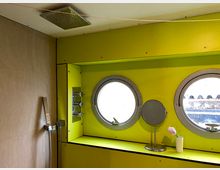 Badezimmer mit ungewöhnlichem Design: Gelbe Wände und runde Bullaugenfenster. Links befinden sich eine Regendusche und Armaturen, rechts ein kleiner Spiegel und eine Vase mit Tulpe auf einer Ablage.
