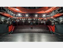 Ein moderner Theatersaal mit Blick von der Bühne auf gestufte Sitzreihen, umgeben von roten Wänden und grauen Akustikelementen. An der Decke befinden sich Scheinwerfer und technische Ausrüstung, während seitliche Treppen den Zugang zu den Sitzplätzen ermöglichen.