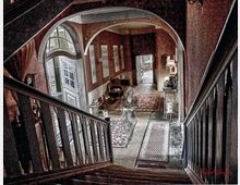 Treppenhaus mit Blick in einen nostalgisch eingerichteten Flur, der mit Teppichen ausgelegt ist. Der Flur hat eine bogenförmige Türöffnung, rote Wände, diverse Möbel wie Tische und Lampen sowie Dekorationen.