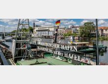Historischer Hafen in Brandenburg an der Havel mit einem alten Schiff und einem Schild, das den Hafenname trägt. Im Hintergrund sind eine Brücke, weitere Boote auf dem Wasser sowie Gebäude und Bäume am Ufer zu sehen.