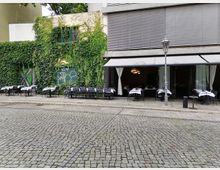 Straßencafé mit Tischen und Stühlen auf einer gepflasterten Fläche vor einem mit Efeu bewachsenen Gebäude. Rechts ist eine offene Fensterfront mit weißen Vorhängen und Markisen zu sehen.