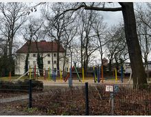Eingezäunter Spielplatz mit Sandfläche, Rutsche und Klettergerüsten aus bunten Pfosten, umgeben von vielen kahlen Bäumen. Im Hintergrund steht ein helles Gebäude mit rotem Ziegeldach sowie weitere Häuser.