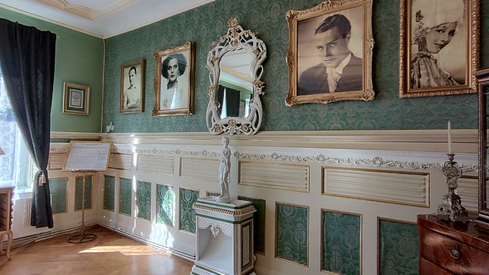 Eleganter Innenraum mit grüner gemusterter Tapete und weißer Wandvertäfelung, geschmückt mit mehreren gerahmten Porträtfotografien und einem reich verzierten Standspiegel. Vor hohen Fenstern mit dunklen Vorhängen stehen ein kleiner Tisch mit Lampe, ein Notenständer sowie eine Kommode mit Kerzenleuchter und verglastem Schaukasten auf Parkettboden.