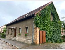 Ein einfaches Wohnhaus mit grauer Fassade und rotem Ziegeldach, von dem eine Seitenwand dicht mit grünem Efeu bewachsen ist. Das Haus hat kleine Fenster mit Blumenkästen und eine Holztür, die über eine kleine Treppe erreichbar ist.