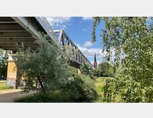 Eine Stahlbrücke mit Gitterstruktur erstreckt sich diagonal über eine grüne Parklandschaft mit Bäumen und Büschen; im Hintergrund ist der Kirchturm einer Backsteinkirche sichtbar. Ein gemauerter Brückenpfeiler zeigt Graffiti, und der Himmel ist leicht bewölkt.