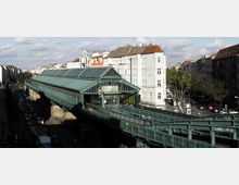 Hochbahnstation in einer städtischen Umgebung mit einer grünen Stahlkonstruktion und Glasdach, flankiert von mehrstöckigen Wohnhäusern und einer belebten Straße mit parkenden Autos auf beiden Seiten.