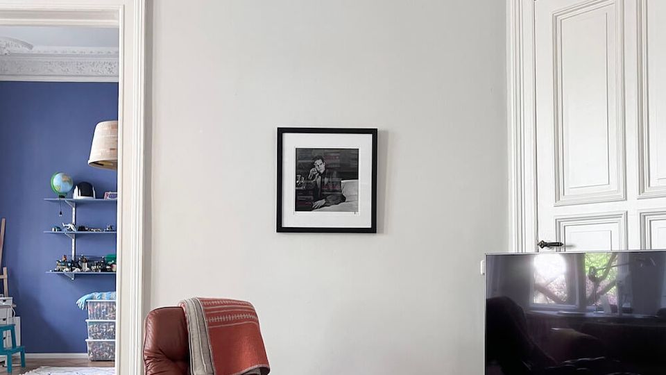 Bright living room with high ceilings, ornate white plaster crown molding, and pale walls, featuring a brown leather lounge chair and a large dark ottoman on a rug. A flat-screen TV sits on a low white media cabinet beside tall white paneled doors, with a wide doorway opening into a blue-walled room with shelving.