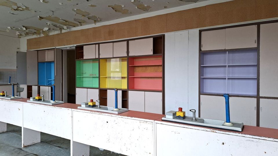 Innenraum eines verlassenen Klassen- oder Laborraums mit langen Arbeitstischen und mehreren Wasserhähnen; an der Rückwand stehen Einbauschränke und Regale mit farbigen Glasfronten. Die Decke zeigt abblätternde Farbe und beschädigten Putz.
