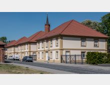 Straßenansicht von mehreren zweigeschossigen Wohnhäusern mit hellen Fassaden und roten Ziegeldächern, im Hintergrund ein spitzer Kirchturm aus rotem Backstein; im Vordergrund geparkte Autos und ein Fußweg mit Straßenlaternen.