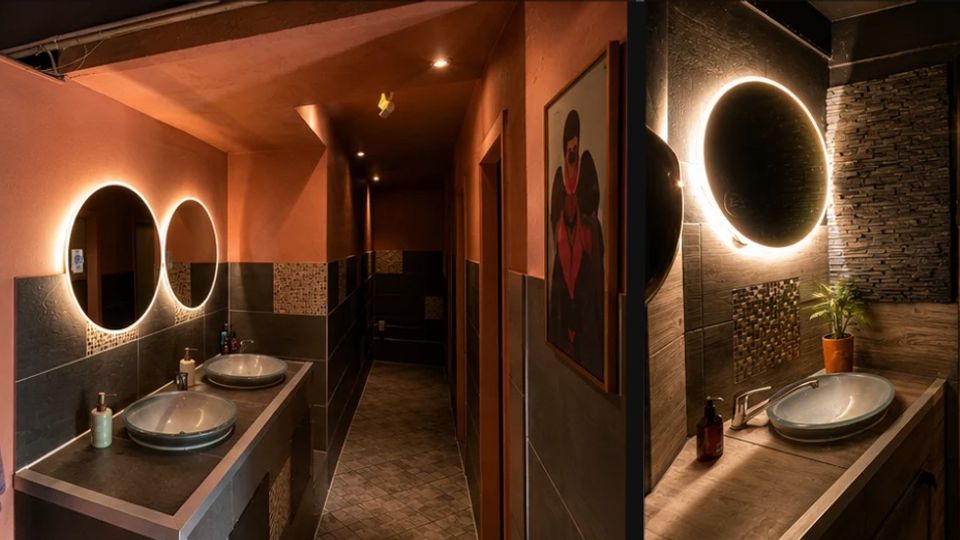 Moderner Waschraum mit zwei runden, hinterleuchteten Spiegeln über Aufsatzwaschbecken auf einer langen Arbeitsplatte, flankiert von rosa Wänden und dunklen Fliesen. Der Bereich führt in einen Flur zu Toilettenkabinen; Akzente setzen strukturierte Steinwandverkleidung, warmes Deckenlicht und ein gerahmtes Wandbild.