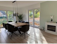 Esszimmer in einem modernen Haus mit einem großen Holztisch und sechs schwarzen Stühlen. Helle, bodentiefe Fenster lassen viel Tageslicht herein und bieten einen Blick auf den herbstlichen Garten; im Raum befinden sich eine grüne Wand, ein dekorativer Kamin und Zimmerpflanzen.