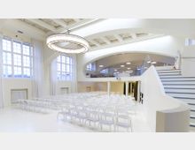 Heller, moderner Veranstaltungsraum mit weißen Wänden, großen Fenstern und einer hohen Decke mit freiliegenden Balken. Der Raum verfügt über Reihen weißer Stühle, eine Freitreppe und einen oberen Bereich mit Glasgeländer.