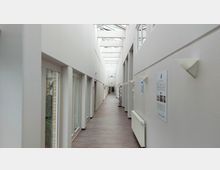 Heller, moderner Flur mit weißen Wänden und einer Decke aus Glasstreben, die Tageslicht hereinlässt. Linke Seite: Türen mit Glasfenstern, rechte Seite: Heizkörper, Wandlampen und Informationsposter; der Boden besteht aus braunem Holz oder Laminat.