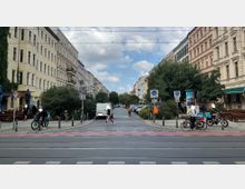 Straßenansicht in einer Wohngegend mit mehrstöckigen Altbauten auf beiden Seiten, einem kleinen Platz mit Bäumen und parkenden Autos in der Mitte. Im Vordergrund befinden sich ein Fahrradweg, Fußgängerüberweg und Schilder für Fahrrad- sowie Parkzonen.