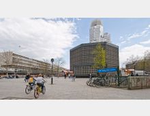 Eine städtische Platzszene mit der Kaiser-Wilhelm-Gedächtniskirche (modernes rechteckiges Gebäude mit markanter Gitterfassade) im Zentrum, flankiert von Wohn- und Geschäftsbauten. Im Vordergrund befinden sich ein U-Bahn-Eingang mit einem Schild "U-Bahnhof Zoologischer Garten" sowie parkende Fahrräder und Passanten.