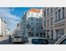 Straßenszene in einer Stadt mit mehrstöckigen Gebäuden im klassischen Baustil. Im Vordergrund befinden sich Fußgänger, ein Fahrradfahrer und parkende Autos; Ampeln und Verkehrsschilder sind sichtbar.