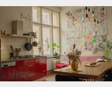 Bright open-plan kitchen and dining area with red lower cabinets, a tiled backsplash, hanging pots, and large multi-pane windows. A dining table sits beside a pink-covered sofa, with potted plants and a large wall collage of colorful drawings under hanging bulb lights.