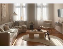 Helles Wohnzimmer mit einer cremefarbenen Eckcouch, einem Holzcouchtisch auf einem beigefarbenen Teppich und einem Sessel mit Schaffell. Im Hintergrund sind große Fenster mit weißen Vorhängen zu sehen, die Tageslicht einlassen, und links hängt eine Wanddekoration aus Stoff.