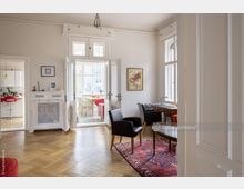 Helles Wohnzimmer mit Parkettboden, einer roten Teppichfläche, schwarzen Sesseln und einem Holztisch. Im Hintergrund führt eine doppelflügelige Glastür in einen Essbereich mit großen Fenstern und einem Tisch mit hellen Stühlen.