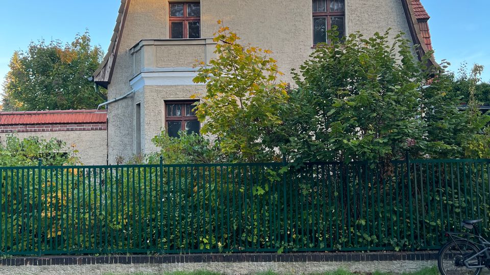 Straßenansicht eines mehrstöckigen Wohnhauses mit heller Putzfassade und steilem Satteldach, teils mit kleinem Balkon und mehreren Fenstern. Vor dem Haus stehen Büsche und Bäume hinter einem grünen Metallzaun; davor verläuft ein Kopfsteinpflasterweg unter blauem Himmel.
